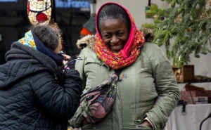 Pascale · 22/11/2025 · 1000 x 618 Cette photographie met en scène deux femmes en interaction dans un marché ou une rue animée. Le sujet principal est la femme en vert, souriante, montrant un objet à une autre personne masquée et vêtue de couleurs vives.La composition est serrée, centrée sur les visages et les mains. Le cadrage est rapproché, créant une ambiance intime. La lumière semble naturelle, douce, avec des ombres peu marquées. Les couleurs dominantes sont les tons chauds (rouge, vert, orange) et les teintes neutres.Le style est documentaire et spontané, évoquant une scène de vie quotidienne. L’ambiance est chaleureuse et conviviale.