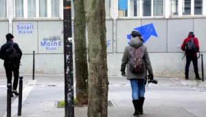 Pascale · 22/11/2025 · 1000 x 569 Cette photographie montre une personne comme sujet principal, vêtue d’un manteau sombre et d’un chapeau, marchant sous un arbre. La composition est simple, avec des éléments urbains en arrière-plan : un panneau "Soleil & Mains", un mur gris et des passants.Le cadrage est serré sur la personne, avec une profondeur de champ réduite. La lumière est naturelle, légèrement tamisée, créant des ombres douces. Les couleurs dominantes sont les tons gris et bleus, avec des touches de vert (l’arbre) et de rouge (vêtements des passants).Le style est réaliste et documentaire, évoquant une ambiance urbaine calme et quotidienne.