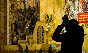 Pascale · 22/11/2025 · 1000 x 603 Cette photographie montre un personnage en silhouette, filmant ou photographiant une fresque murale. Le sujet principal est cette œuvre représentant deux hommes en uniforme, entourée d'éléments architecturaux stylisés.La composition est verticale, avec un cadrage serré sur le mur décoré. La lumière est tamisée, créant des contrastes doux. Les couleurs dominantes sont des tons chauds (jaunes, ocres) et des touches de vert.Le style évoque un réalisme socialiste, typique des fresques historiques. L'ambiance est à la fois solennelle et mystérieuse, renforcée par l'ombre du photographe.