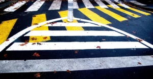 Franck T. · 08/11/2025 · 1000 x 521 Cette photographie met en scène un **passage piéton** partiellement endommagé, avec des bandes blanches et jaunes effacées ou déformées. Le **sujet principal** est la dégradation des marquages au sol, soulignant leur usure.La **composition** est centrée, avec un **cadrage serré** qui isole les lignes du passage. La **lumière**, naturelle et tamisée, crée des ombres douces, accentuant les contrastes. Les **couleurs dominantes** sont le **noir** (asphalte), le **blanc** et le **jaune** des marquages, ainsi que des touches de **rouge et marron** (feuilles mortes).Le style est **réaliste et documentaire**, évoquant une **ambiance urbaine négligée**, presque mélancolique.