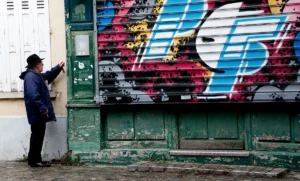 Franck T. · 08/11/2025 · 1200 x 723 Cette photographie montre un homme en train de peindre ou d’inspecter un graffiti sur un mur vert délabré. Le sujet principal est ce mur couvert de tags colorés, dont un visage stylisé en bleu et rouge domine.La composition est serrée, avec un cadrage en plan moyen. La lumière semble naturelle, un peu grise, typique d’un jour couvert. Les couleurs dominantes sont les teintes vives des graffitis contrastant avec le vert usé du mur.Le style est urbain et spontané, évoquant une ambiance de rue authentique et artistique, mêlant créativité et négligence.