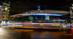 Cette photographie capture une scène urbaine nocturne dynamique. Le sujet principal est un bâtiment moderne aux lignes arrondies, éclairé par des néons bleus. La composition met en valeur des traînées lumineuses, créées par le mouvement des véhicules, suggérant une exposition longue. Le cadrage est large, incluant une partie de la route et des bâtiments environnants.Les couleurs dominantes sont les bleus froids, les tons chauds des phares et les éclats de lumière blanche. Le style est photographique moderne, avec un effet artistique grâce aux traînées lumineuses. L’ambiance est vibrante et énergique, reflétant l’agitation nocturne d’une ville.