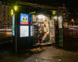 Cette photographie montre un kiosque à journaux ouvert la nuit, éclairé par une lumière artificielle chaude. Le sujet principal est le vendeur, partiellement flou, derrière les étagères remplies de magazines et produits divers.La composition est symétrique, avec le kiosque au centre. Le cadrage est serré, mettant en valeur les détails intérieurs et extérieurs. Les couleurs dominantes sont des tons verts, jaunes et blancs, créant une ambiance nocturne et chaleureuse. Le style est réaliste et légèrement artistique grâce aux effets de flou et de lumière. L’ambiance est intimiste et vivante, malgré l’heure tardive.