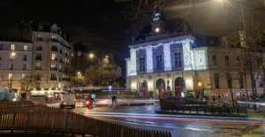 Cette photographie capture une place urbaine nocturne. Le sujet principal est un bâtiment historique éclairé en bleu, probablement un théâtre ou une mairie. La composition montre une perspective large avec des bâtiments résidentiels autour et une rue légèrement courbée.Le cadrage inclut des éléments architecturaux et des passants floutés par le mouvement. Les lumières artificielles dominent, créant des contrastes nets entre ombres et éclairages. Les couleurs sont principalement bleutées, grises et jaunâtres, renforçant une ambiance nocturne et mystérieuse. Le style est réaliste et photographique, évoquant une atmosphère calme et légèrement mélancolique.