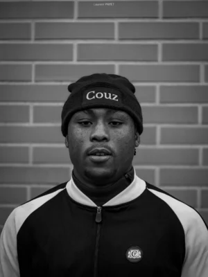 Laurent · 2160 x 2877 Cette photographie met en scène un jeune homme comme sujet principal, posé devant un mur en briques. Le cadrage est serré, centré sur son visage et sa partie supérieure du torse. Il porte un bonnet et une veste sport à capuche, avec des détails contrastés.La lumière est douce mais contrastée, créant des ombres marquées, typiques d’un éclairage studio. Les couleurs dominantes sont le noir et blanc, donnant un style sobre et moderne. L’ambiance est à la fois sérieuse et introspective, renforcée par le fond neutre et le jeu d’ombres.