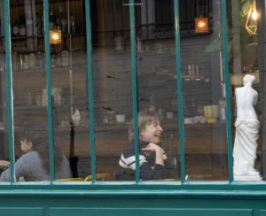 Laurent · 2657 x 2160 Cette photographie montre un sujet principal flou, une personne assise à l’intérieur d’un café ou d’un restaurant, visible à travers une vitre striée. La composition joue sur les reflets et les ombres, créant une ambiance intimiste et légèrement mélancolique.Le cadrage est serré, mettant en valeur la vitre et les détails intérieurs. La lumière est douce et tamisée, avec des teintes chaudes provenant des lampes. Les couleurs dominantes sont des tons neutres (beiges, gris) et des touches de bleu et de vert.Le style est réaliste et documentaire, évoquant une scène de vie quotidienne. L’ambiance est calme et légèrement nostalgique.