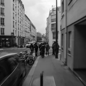 Laurent · 2160 x 2160 Cette photographie en noir et blanc capture une rue urbaine animée. Le sujet principal est un groupe de personnes marchant sur un trottoir étroit, encadré par des bâtiments résidentiels et commerciaux.La composition est dynamique, avec une perspective légèrement inclinée et une profondeur de champ réduite, mettant l'accent sur les passants. Le cadrage est serré, centré sur les piétons et les véhicules en mouvement. La lumière est diffuse, typique d'un jour couvert, adoucissant les contrastes.Les tons dominants sont les gris et noirs, renforçant l'ambiance moderne et légèrement mélancolique. Le style est documentaire et réaliste, évoquant une atmosphère quotidienne et urbaine.