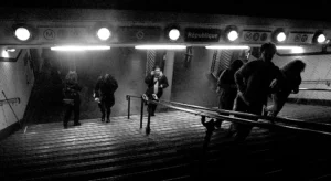 Cette photographie en noir et blanc capture une scène de gare ou de quai souterrain. Le sujet principal semble être un groupe de personnes en mouvement, légèrement floues, suggérant une ambiance dynamique et pressée. La composition utilise une perspective descendante, avec des lignes directrices (escaliers, rails) convergeant vers le fond.La lumière est douce mais contrastée, éclairant les visages et créant des ombres marquées. Les couleurs dominantes, bien sûr, sont les nuances de gris, renforçant l'atmosphère dramatique et intemporelle. Le style rappelle le cinéma classique, évoquant une ambiance mélancolique et mystérieuse.