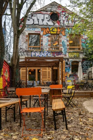 Antoine · 22/11/2025 · 1667 x 2500 Cette photographie montre un petit café extérieur nommé "STR" décoré de graffitis colorés. Le sujet principal est l’établissement, avec ses murs couverts d’art urbain et ses tables en bois disposées devant.Le cadrage inclut des arbres et une ambiance naturelle. La lumière est douce, probablement naturelle, créant des ombres légères. Les couleurs dominantes sont vives et variées, avec des tons de bleu, rouge, jaune et vert.Le style est bohème et artistique, évoquant une ambiance décontractée et créative, typique d’un lieu alternatif et convivial.