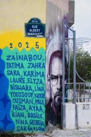 Antoine · 22/11/2025 · 2000 x 3000 Cette photographie montre un mur tagué avec un portrait en noir et blanc d’un homme, entouré de noms de femmes écrites en bleu : "Zainabou, Fatima Zahra...". Le cadrage est serré sur le panneau et inclut une partie d’un bâtiment jaune en arrière-plan.La lumière semble naturelle, avec des contrastes marqués. Les couleurs dominantes sont le bleu des inscriptions, le noir et blanc du portrait, et le jaune vif du mur adjacent. Le style est urbain et artistique, évoquant une ambiance militante ou commémorative, typique des fresques engagées.