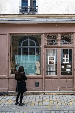 Antoine · 22/11/2025 · 1667 x 2500 Cette photographie met en scène une femme en manteau noir, sujet principal, qui semble observer une vitrine. La composition est simple, avec un cadrage serré sur la porte et la vitrine colorée en bleu-vert. La lumière naturelle éclaire la scène, douce et légèrement contrastée. Les couleurs dominantes sont les tons pastel (rose, bleu) du bâtiment et le noir du manteau de la femme.L’ambiance est calme et urbaine, évoquant une rue paisible. Le style est réaliste et documentaire, avec une attention portée aux détails architecturaux et à l’atmosphère quotidienne.