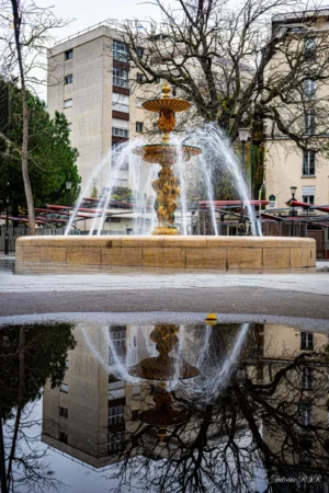 Antoine · 22/11/2025 · 2000 x 3000 Cette photographie met en scène une **fontaine monumentale** comme sujet principal, avec des jeux d’eau et une structure ornée. La **composition** est symétrique, renforcée par le reflet parfait dans l’eau, créant une impression de miroir. Le **cadrage** est serré sur la fontaine, avec des bâtiments et des arbres en arrière-plan.La **lumière** semble naturelle, douce, probablement en journée, avec des ombres discrètes. Les **couleurs dominantes** sont des tons neutres (beige, gris) et des reflets dorés de la fontaine. Le style est **classique et élégant**, évoquant une ambiance **calme et intemporelle**, presque urbaine et poétique.