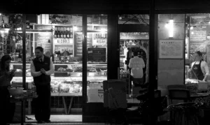Pascale · 20/12/2025 · 1000 x 596 Cette photographie en noir et blanc capture une scène nocturne devant un magasin éclairé. Le sujet principal semble être les deux personnes à l’intérieur, dont une est derrière un comptoir rempli de produits alimentaires. La composition met en valeur les vitrines et les enseignes lumineuses à travers les fenêtres.Le cadrage est serré sur les vitrines, créant une ambiance intimiste. La lumière, bien que tamisée, éclaire les produits et les personnes, contrastant avec l’obscurité extérieure. Les couleurs dominantes sont les tons de gris, typiques du noir et blanc, renforçant une ambiance rétro et nostalgique. Le style est documentaire et réaliste.