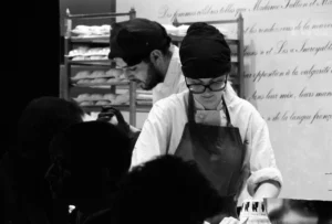 Pascale · 20/12/2025 · 5088 x 3440 Cette photographie en noir et blanc montre deux personnes en cuisine, probablement des chefs. Le sujet principal semble être le chef de droite, en train de travailler.La composition met en avant un cadrage serré sur les individus, avec des éléments de fond flous (étagères, textes). La lumière est douce, éclairant les sujets de manière naturelle, sans ombres marquées. Les couleurs dominantes sont les nuances de gris typiques du noir et blanc.Le style est documentaire et réaliste, capturant un moment authentique. L’ambiance est professionnelle et concentrée, évoquant un environnement de travail intense et spécialisé.