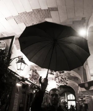 Pascale · 20/12/2025 · 840 x 1000 Cette photographie en noir et blanc met en scène un **parapluie noir** comme sujet principal, ouvert et dominant le cadrage. La composition joue sur les lignes architecturales et les perspectives, avec des éléments urbains comme des enseignes et des lanternes.La lumière, douce et tamisée, crée des contrastes entre ombres et clartés, accentuant l’ambiance mystérieuse et poétique. Les tons gris et noirs dominent, renforçant l’aspect dramatique et intemporel. Le style rappelle le photojournalisme classique ou une esthétique vintage, évoquant une atmosphère mélancolique et contemplative dans un cadre urbain.