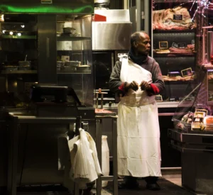 Pascale · 20/12/2025 · 4160 x 3816 Cette photographie montre un cuisinier comme sujet principal, vêtu d’une tenue blanche et de gants rouges, en train de travailler dans une cuisine professionnelle. La composition met en valeur son activité au centre, entourée d’équipements culinaires et d’ingrédients.Le cadrage est serré, capturant une ambiance réaliste et dynamique. La lumière est tamisée mais efficace, éclairant les zones de travail. Les couleurs dominantes sont les tons chauds (rouge des gants, blanc de la tenue) et les teintes industrielles (métalliques, vert des néons).Le style est documentaire, reflétant une ambiance authentique et intense de la vie en cuisine.