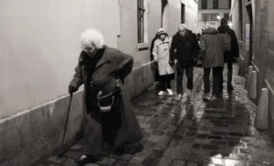 Pascale · 20/12/2025 · 1000 x 606 Cette photographie en noir et blanc montre un vieil homme comme sujet principal, penché pour ramasser quelque chose dans une ruelle étroite. La composition met en valeur cet individu au premier plan, tandis que d’autres personnes, floues, déambulent en arrière-plan.Le cadrage serré accentue l’intimité et la solitude du personnage. La lumière, tamisée, crée des contrastes doux, typiques d’une ambiance nocturne ou crépusculaire. Les tons dominants, gris et noirs, renforcent une atmosphère mélancolique et sobre. Le style, réaliste et documentaire, évoque une scène de vie quotidienne, imprégnée de nostalgie et de simplicité.
