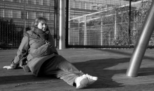 Pascale · 06/12/2025 · 1250 x 743 Cette photographie en noir et blanc met en scène un homme comme sujet principal, assis par terre, les jambes étendues, dans une posture décontractée. La composition joue sur les lignes architecturales avec une structure métallique en arrière-plan. Le cadrage est serré sur le sujet, avec une légère inclinaison vers lui.La lumière semble naturelle, douce et diffuse, créant des contrastes modérés. Les tons dominants sont les gris et les noirs, typiques du noir et blanc, renforçant une ambiance contemplative et mélancolique. Le style est documentaire et intime, évoquant une atmosphère calme et réfléchie.
