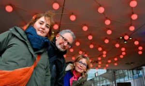 Pascale · 06/12/2025 · 1000 x 596 Cette photographie montre trois personnes souriantes posant sous une structure lumineuse. Le sujet principal est leur joie et complicité.La composition est symétrique, avec un cadrage serré sur les visages. La lumière est douce et artificielle, provenant des sphères lumineuses rouges en arrière-plan. Les couleurs dominantes sont les tons chauds (rouge, orange) et les teintes neutres des vêtements.Le style est naturel et spontané, évoquant une ambiance conviviale et festive. L’éclairage et les lumières créent une atmosphère chaleureuse et joyeuse.