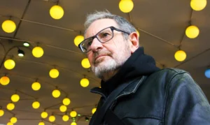Pascale · 06/12/2025 · 1250 x 744 Cette photographie met en scène un homme d'âge mûr comme sujet principal, vêtu d'une veste sombre et portant des lunettes. Le cadrage est serré sur son visage, créant une ambiance intime. L'arrière-plan, composé de multiples boules lumineuses jaunes, apporte une touche décorative et festive.La lumière est douce et uniforme, éclairant bien le visage tout en mettant en valeur les teintes chaudes des boules. Les couleurs dominantes sont les tons jaunes vifs et les noirs/gris de la veste. Le style est sobre et naturel, évoquant une ambiance à la fois moderne et chaleureuse.