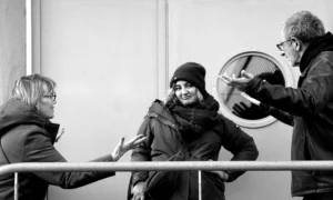 Pascale · 06/12/2025 · 1500 x 902 Cette photographie en noir et blanc montre trois personnes comme sujets principaux : une femme au centre, souriante, et deux hommes de chaque côté, l’un lui tendant un objet.La composition est dynamique, centrée sur l’interaction entre les personnages. Le cadrage est serré, mettant l’accent sur leurs visages et gestes. La lumière est douce et naturelle, éclairant uniformément la scène sans ombres marquées.Le style est réaliste et spontané, évoquant une ambiance chaleureuse et conviviale, presque théâtrale. Les tons gris dominent, renforçant l’aspect intemporel de l’image.