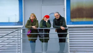 Pascale · 06/12/2025 · 1000 x 578 Cette photographie montre trois personnes debout sur une passerelle métallique. Le sujet principal est le trio, composé de deux femmes et d’un homme, tous vêtus chaudement, avec des expressions neutres ou légèrement souriantes.La composition est symétrique, avec les personnages alignés sur la rampe. Le cadrage est serré, centré sur eux, avec un arrière-plan flou de paysage hivernal. La lumière est naturelle, douce mais froide, typique d’un jour d’hiver. Les couleurs dominantes sont les tons neutres (gris, bleu pâle) et quelques touches de vert et rouge dans leurs vêtements. L’ambiance est calme et contemplative, évoquant une pause dans un voyage ou une réflexion.