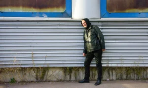 Pascale · 06/12/2025 · 1250 x 742 Cette photographie met en scène un homme comme sujet principal, vêtu d’une veste verte et d’un pantalon noir, marchant près d’un mur métallique dégradé. La composition est simple et épurée, avec un cadrage serré qui isole le personnage.La lumière semble naturelle, légèrement tamisée, créant des ombres douces. Les couleurs dominantes sont les tons froids du métal rouillé (bleu-gris) et les teintes sombres du vêtement de l’homme, contrastant avec sa veste verte.Le style est réaliste et sobre, évoquant une ambiance urbaine et légèrement mélancolique.