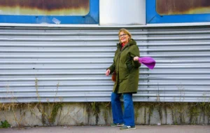 Pascale · 06/12/2025 · 1000 x 633 Cette photographie met en scène une femme comme sujet principal, vêtue d’un manteau vert et tenant un objet rose. La composition est simple et épurée, avec la femme positionnée devant un mur gris strié, légèrement incliné.Le cadrage est serré, centré sur elle, avec un arrière-plan minimaliste. La lumière semble naturelle, douce et légèrement diffuse, créant des ombres peu marquées. Les couleurs dominantes sont les tons verts du manteau, gris du mur et rose vif de l’objet tenu.Le style est réaliste et direct, évoquant une ambiance sobre et contemporaine, presque intimiste.