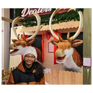 Cette photographie montre une personne souriante posant devant deux grandes décorations en forme de têtes de rennes. Le sujet principal est cette personne, vêtue d’un manteau brun et d’un bonnet à cornes.La composition est symétrique, avec les têtes de rennes de chaque côté. Le cadrage est serré, mettant en valeur le visage et les décorations. La lumière semble naturelle et douce, éclairant bien les sujets.Les couleurs dominantes sont les tons bruns et blancs des rennes, avec des touches de rouge et vert en arrière-plan. L’ambiance est festive et chaleureuse, évoquant les fêtes de fin d’année.