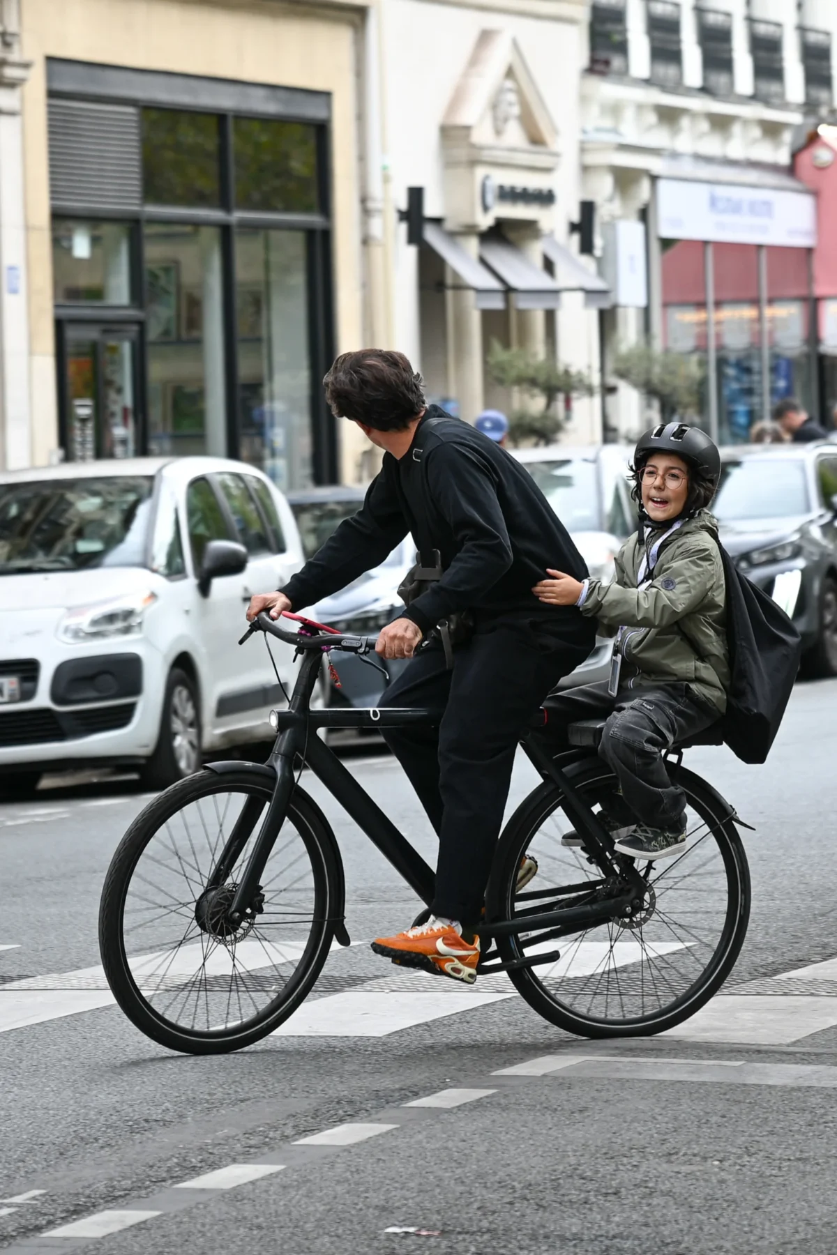Jean-Marc · 13/09/2025 · 1925 x 2888 Cette photographie montre deux personnes à vélo en ville. Le sujet principal est un adulte en costume poussant un vélo avec un enfant assis devant, portant un casque et une veste verte.La composition est dynamique, avec un cadrage serré sur les cyclistes. L'arrière-plan montre une rue urbaine avec des voitures et des bâtiments. La lumière est naturelle, probablement en journée, avec des ombres nettes. Les couleurs dominantes sont les tons neutres (gris, noir) et les couleurs vives comme le vert de la veste de l'enfant.L'ambiance est urbaine et quotidienne, évoquant un moment de partage et de mobilité en ville.