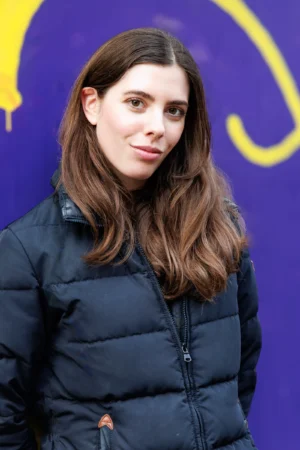 Claude · 2883 x 4324 Cette photographie met en scène une jeune femme comme sujet principal, cadrée en plan serré. Elle porte un manteau noir mat et a les cheveux longs et ondulés.La composition est simple, centrée sur son visage et son haut du corps. La lumière semble naturelle, douce et légèrement latérale, éclairant son visage sans ombres marquées.Les couleurs dominantes sont le noir de son manteau et les tons froids du fond bleu-violet. L’ambiance est sobre et contemporaine, avec une touche de modernité et de neutralité.