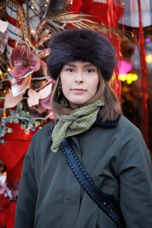 Claude · 4000 x 6000 Cette photographie met en scène une jeune femme comme sujet principal, vêtue d’un manteau vert foncé, d’un foulard et d’un chapeau en fourrure. Le cadrage est serré, centré sur elle, avec un arrière-plan flou décoré de guirlandes et de décorations festives colorées.La lumière est douce et naturelle, éclairant son visage avec une ambiance chaleureuse. Les couleurs dominantes sont les tons verts, bruns et rouges, renforçant une ambiance hivernale et festive. Le style est sobre et élégant, évoquant une touche urbaine et contemporaine. L’ambiance générale est conviviale et joyeuse, typique d’une période de fêtes.