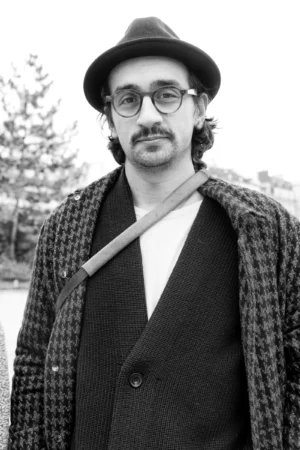 Claude · 4000 x 6000 Cette photographie en noir et blanc met en scène un homme comme sujet principal, vêtu d’un veston à carreaux, d’un pull et d’une cravate, avec des lunettes rondes. Le cadrage est serré, centré sur lui, avec un fond flou de végétation.La lumière est douce et naturelle, sans contraste marqué. L’ambiance est sobre et introspective, évoquant une touche rétro ou artistique. Les tons dominants, en niveaux de gris, renforcent un style classique et élégant, avec une atmosphère calme et réfléchie.