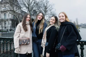 Claude · 6000 x 4000 Cette photographie montre quatre jeunes femmes souriantes posant ensemble. Le sujet principal est leur complicité et leur harmonie.La composition est symétrique, avec les quatre femmes alignées côte à côte. Le cadrage est serré sur elles, avec un arrière-plan flou de bâtiments et de végétation.La lumière semble naturelle, légèrement douce, typique d’un jour nuageux. Les couleurs dominantes sont les tons neutres (beige, noir) et quelques touches de couleurs vives (veste rose et accessoires).Le style est décontracté et moderne, évoquant une ambiance amicale et chaleureuse.