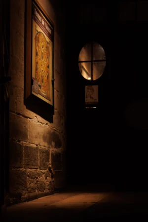 Claude · 4000 x 6000 Cette photographie met en scène un couloir sombre et étroit. Le sujet principal est une lumière tamisée éclairant une partie du mur en pierre, révélant une texture rugueuse. Un cadre doré, probablement une œuvre ou un miroir, et une boule lumineuse fixée au mur sont visibles.La composition est minimaliste, avec un cadrage serré qui accentue l'effet de profondeur et de mystère. La lumière, douce et contrastée, crée une ambiance dramatique. Les couleurs dominantes sont les tons chauds (jaunes, ors) et les teintes sombres (noirs, gris). Le style évoque une atmosphère intemporelle et presque cinématographique, renforçant une impression de solitude et de mélancolie.