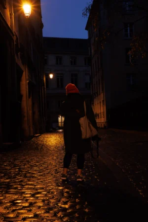 Claude · 3782 x 5673 Cette photographie capture une personne en mouvement dans une ruelle étroite et pavée, la nuit. Le sujet principal, vêtu d’une veste sombre et d’un bonnet rouge, est flou, suggérant un déplacement rapide.La composition est dynamique, avec un cadrage serré qui guide le regard vers le centre. Les murs anciens et les bâtiments encadrants créent une ambiance intimiste. La lumière, douce et tamisée, provient d’une lampe à l’extrémité gauche, éclairant partiellement la scène.Les couleurs dominantes sont les tons sombres (noirs, gris) avec une touche de rouge vif. Le style est réaliste et légèrement dramatique, renforçant une ambiance mystérieuse et poétique.