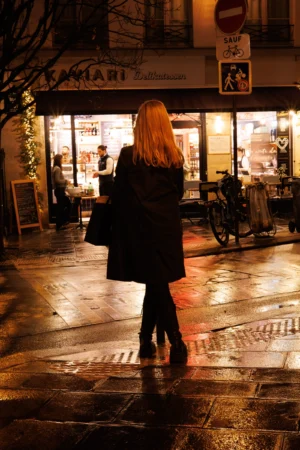 Claude · 3463 x 5194 Cette photographie met en scène une femme au premier plan, vue de dos, portant un manteau noir et tenant un sac. La composition joue sur la profondeur avec un restaurant éclairé en arrière-plan.Le cadrage est serré sur la silhouette, créant un effet de flou artistique. La lumière est douce et tamisée, avec des éclairages chauds provenant des vitrines et des lampadaires, soulignant une ambiance nocturne et intimiste. Les couleurs dominantes sont les tons sombres (noir, gris) adoucis par des éclats dorés.Le style est réaliste et légèrement poétique, évoquant une scène urbaine paisible et mélancolique.
