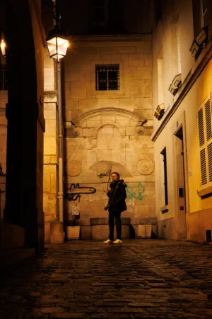 Claude · 3821 x 5731 Cette photographie met en scène une personne comme sujet principal, debout dans une ruelle étroite, éclairée par une lumière douce. La composition joue sur les lignes architecturales convergentes et le cadrage serré, créant une ambiance intimiste.Les tons dominants sont des jaunes chauds et des ombres profondes, renforçant l'atmosphère nocturne. La lumière, tamisée et légèrement dorée, provient probablement d'un réverbère, ajoutant une touche mélancolique et poétique. Le style évoque un réalisme artistique, avec une ambiance contemplative et presque cinématographique.