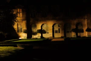Claude · 5764 x 3843 Cette photographie met en scène un bâtiment historique, probablement un château ou une demeure ancienne, éclairé la nuit. Le sujet principal est l’architecture majestueuse avec ses fenêtres et ses arcs.La composition repose sur un cadrage serré, mettant en valeur les jeux d’ombres et de lumières. Les arbres taillés encadrent l’entrée, ajoutant une touche de symétrie.Les couleurs dominantes sont les tons sombres et les éclairages chauds jaunâtres, créant une ambiance mystérieuse et élégante. Le style est photographique classique, avec une ambiance nocturne dramatique et poétique.