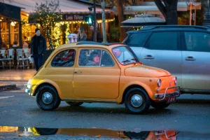 Jean-Marc · 20/12/2025 · 5070 x 3380 Cette photographie met en scène une **Fiat 500 vintage jaune** comme sujet principal. La composition est dynamique, avec un cadrage serré sur la voiture, légèrement de trois-quarts, reflétée sur une flaque d’eau.La lumière douce, probablement crépusculaire, crée une ambiance chaleureuse et tamisée. Les couleurs dominantes sont le **jaune vif** de la voiture, contrastant avec les tons **bleutés et gris** de l’arrière-plan urbain.Le style évoque un mélange de nostalgie et de modernité, renforcé par les éléments architecturaux et les passants en arrière-plan. L’ambiance est à la fois **romantique et vivante**.