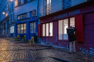 Jean-Marc · 20/12/2025 · 5247 x 3498 Cette photographie capture une rue nocturne pittoresque. Le sujet principal est un homme en imperméable, de dos, marchant près d’un bâtiment éclairé. La composition met en valeur une galerie d’art nommée "La Galerie Rouge", avec ses vitrines lumineuses et ses couleurs vives.Le cadrage est serré, centré sur le passage étroit et les façades colorées. La lumière douce, bleutée et artificielle, crée une ambiance tamisée et chaleureuse. Les couleurs dominantes sont les tons bleus, rouges et blancs, renforçant l’aspect artistique et urbain. Le style est réaliste et photographique, évoquant une ambiance poétique et mélancolique, typique des scènes nocturnes en ville.