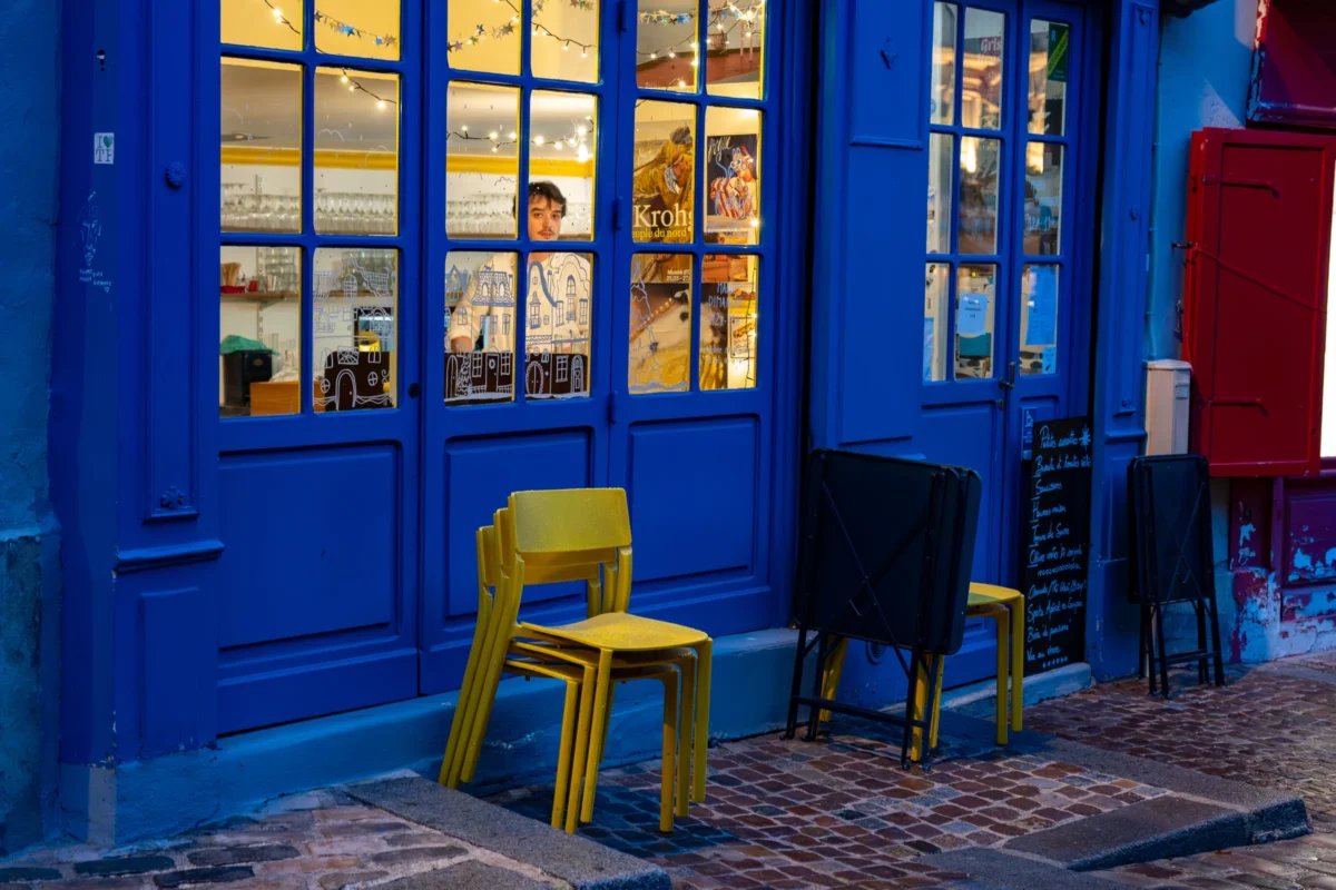 Jean-Marc · 20/12/2025 · 4852 x 3235 Cette photographie met en scène une entrée colorée aux portes bleues et vitres éclairées. Le sujet principal est l’espace extérieur avec deux chaises, l’une jaune et l’autre noire, placées sur un sol pavé.La composition est symétrique et simple, avec un cadrage serré sur l’entrée. La lumière, douce et tamisée, provient de l’intérieur, créant une ambiance chaleureuse et intimiste. Les couleurs dominantes sont le bleu vif des portes et le jaune éclatant de la chaise, contrastant avec le noir et le gris des autres éléments.Le style évoque un mélange de modernité et de simplicité, tandis que l’ambiance est à la fois accueillante et légèrement mystérieuse.