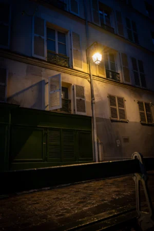 Jean-Marc · 20/12/2025 · 3451 x 5176 Cette photographie capture une rue nocturne urbaine. Le sujet principal est un bâtiment ancien éclairé par un réverbère, dont la lumière jaunâtre crée un contraste avec l'obscurité ambiante. La composition met en valeur les fenêtres éclairées et les ombres marquées, renforçant l'atmosphère mystérieuse.Le cadrage serré et légèrement bas accentue la verticalité du bâtiment et du réverbère. Les couleurs dominantes sont les tons froids (noirs et gris) contrastant avec la lumière chaude du réverbère. Le style est réaliste et photographique, évoquant une ambiance mélancolique et poétique, typique des scènes urbaines nocturnes.