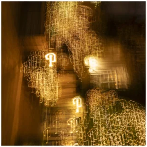 Cette photographie capture une structure métallique complexe, probablement une sculpture ou une installation lumineuse. Le sujet principal semble être les jeux de lumière et d'ombres créant des effets dynamiques.La composition est centrée, avec un cadrage serré qui accentue les détails des motifs géométriques. La lumière, douce et diffuse, produit des halos lumineux et des traînées floues, évoquant un mouvement. Les couleurs dominantes sont des tons chauds : oranges, jaunes et bruns, renforçant une ambiance mystérieuse et onirique. Le style rappelle une approche artistique abstraite et expérimentale.