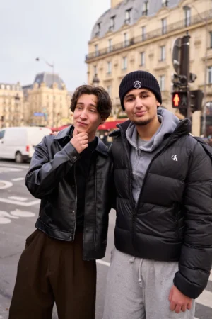 Gief · 1080 x 1620 Cette photographie montre deux jeunes hommes posant côte à côte dans une rue urbaine. Le sujet principal est leur interaction amicale, l’un tenant un cornet de glace.La composition est symétrique et serrée, avec un cadrage en plan serré. La lumière est naturelle, légèrement diffuse, typique d’un jour nuageux. Les couleurs dominantes sont les tons neutres (gris, noir) des vêtements et du décor, adoucis par les touches de rouge (casquette) et de blanc (glace).Le style est spontané et décontracté, évoquant une ambiance jeune et détendue dans un cadre citadin.