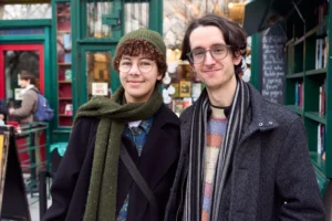 Gief · 2430 x 1620 Cette photographie met en scène deux jeunes hommes comme sujets principaux, posant côte à côte avec des sourires discrets. Le cadrage est serré sur eux, avec un arrière-plan flou montrant des vitrines colorées et des enseignes.La lumière est naturelle et douce, probablement en journée. Les couleurs dominantes sont les tons neutres (noirs, gris, marron) de leurs vêtements, contrastés par des touches de vert et de rouge dans l’arrière-plan. Le style est décontracté et urbain, évoquant une ambiance chaleureuse et conviviale dans un quartier animé.