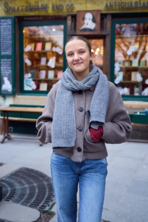 Gief · 1080 x 1620 Cette photographie met en scène une jeune femme comme sujet principal, souriante et détendue. Elle porte un style décontracté avec un gilet texturé, un foulard et un jean.La composition est simple et naturelle, centrée sur elle. Le cadrage est serré, incluant partiellement un magasin d'antiquités en arrière-plan.La lumière semble douce et naturelle, probablement en journée. Les couleurs dominantes sont les tons neutres (gris, bleu) avec des touches de rouge (gobelet).L’ambiance est chaleureuse et conviviale, évoquant une journée urbaine paisible.