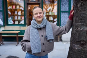 Gief · 2430 x 1620 Cette photographie met en scène une jeune femme comme sujet principal, souriante et détendue, vêtue d’un pull beige et d’un foulard gris. Le cadrage est serré sur elle, avec un arrière-plan flou montrant une façade de bâtiment et des tables à l’extérieur.La lumière est naturelle et douce, probablement en journée. Les couleurs dominantes sont les tons neutres (beige, gris) et quelques touches de rouge et de vert. Le style est simple et naturel, évoquant une ambiance chaleureuse et décontractée, presque urbaine et quotidienne.