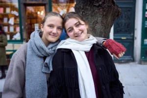 Gief · 2430 x 1620 Cette photographie met en scène deux jeunes femmes souriantes posant côte à côte. Le sujet principal est leur complicité et leur joie apparente.La composition est simple et symétrique, avec un cadrage serré sur elles. L’arrière-plan montre un arbre et une façade urbaine, légèrement flou. La lumière naturelle, douce et légèrement tamisée, crée une ambiance chaleureuse. Les couleurs dominantes sont les tons neutres (gris, beige) et les touches de rouge et blanc apportées par leurs vêtements. Le style est naturel et spontané, évoquant une ambiance conviviale et authentique.