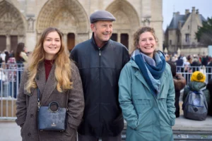 Gief · 2430 x 1620 Cette photographie montre trois personnes marchant devant une cathédrale gothique. Le sujet principal est ce trio souriant, composé de deux femmes et d’un homme, habillés en vêtements chauds et décontractés.La composition est équilibrée, avec un cadrage serré sur les trois individus. La lumière est naturelle et douce, suggérant une journée claire. Les couleurs dominantes sont les tons neutres (beige, gris) des vêtements et bâtiments, adoucis par des touches de vert et de marron.Le style est documentaire et chaleureux, évoquant une ambiance conviviale et touristique.